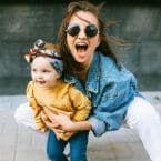 A joyful mother, wearing sunglasses and a denim jacket, squats and holds her toddler daughter, both smiling and having fun. The little girl wears a yellow shirt and a headband.