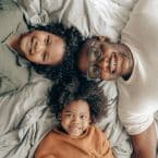 Smiling Black family lies on gray bedspread. Father wears glasses, children have curly hair. Warm, loving moment. Considering roof cleaning for a safe home.