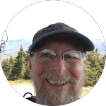 Smiling man with a beard and glasses wearing a hat, with a mountainous, tree-covered landscape in the background.