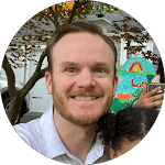Smiling man with a beard poses outdoors with a child near a colorful painted sign. The background shows a house with a garage.