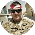 Air Force member in uniform, wearing sunglasses and a red bandana, stands in a desert setting. The uniform has "U.S. AIR FORCE" on it.