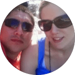A couple wearing sunglasses poses for a selfie on a sunny day, possibly at the beach. The man wears a red shirt, and a blue object is visible between them.